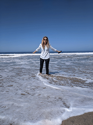 Kristin is pictured in a light blue jean shirts and black pants standing on the beach in the ocean with arms outward.