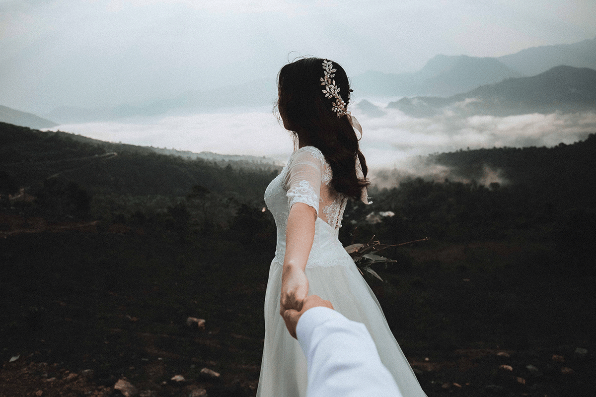 A woman in a white wedding dress with dark hair is facing misty mountains. She has a beaded tiara in the pattern of leaves. The groom (out of the frame) is holding her hand as if pulling her.