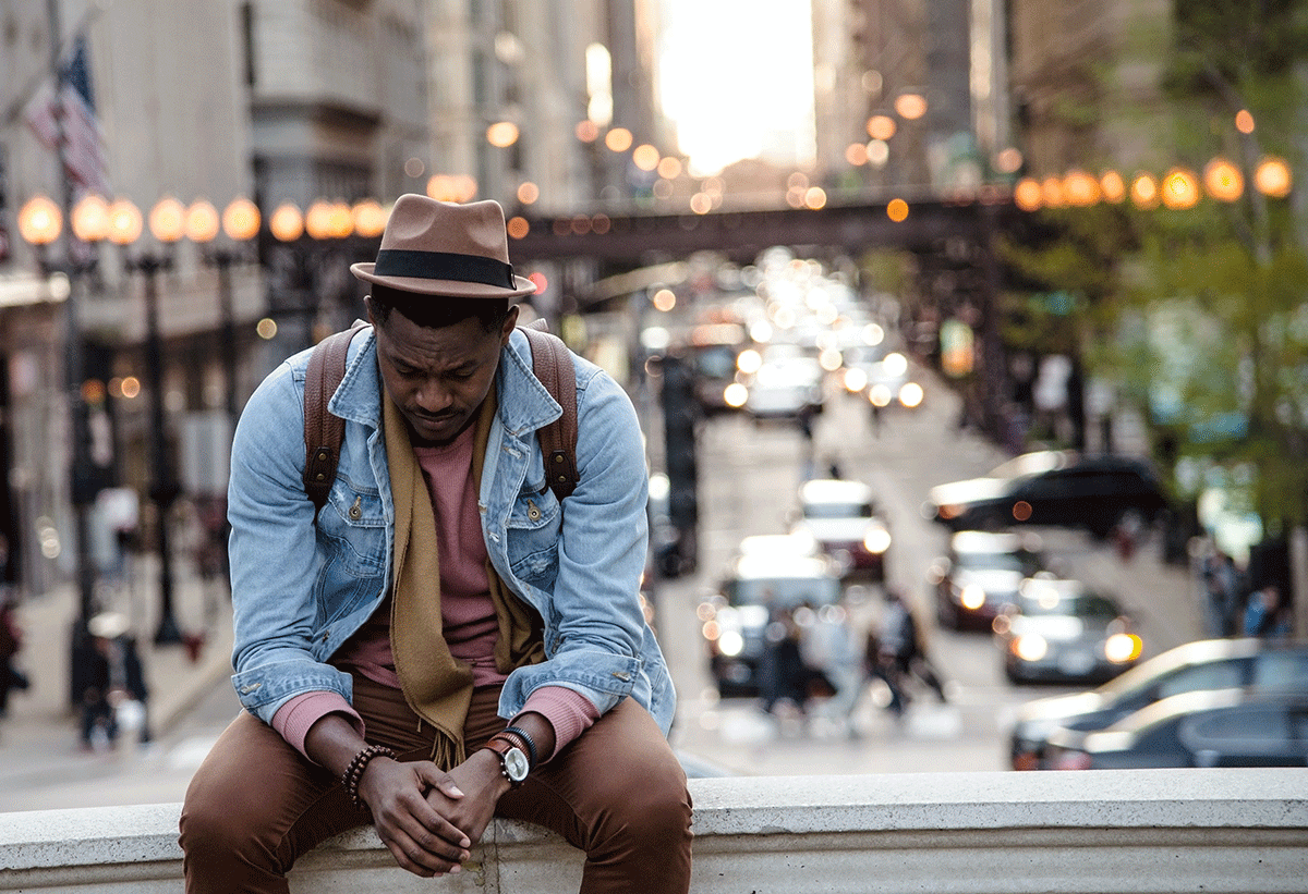 A man dressed in brown pants and a brown t-shirt, jean jacket and brown hat sits on a bridge with hands folded eyes fixed on the ground.