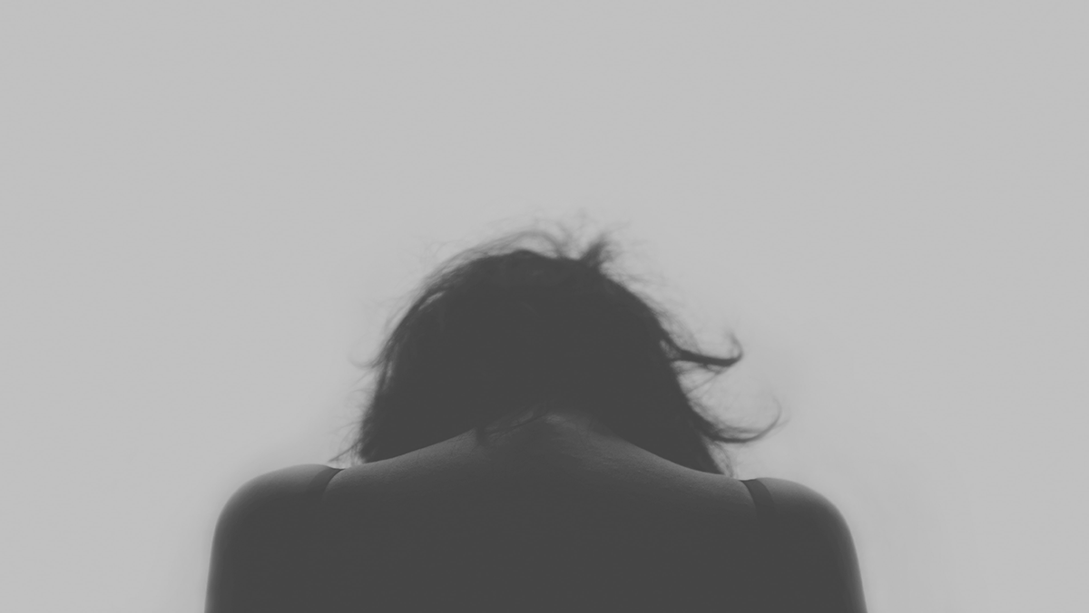 A black and white photo of a woman with her back turned towards the viewer and head looking down towards the ground.