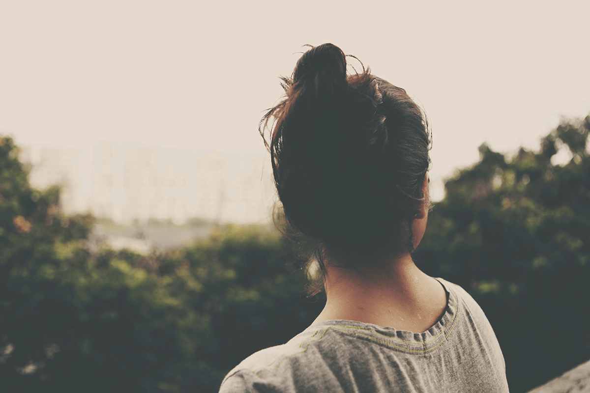 A woman wiA woman with brown hair swept up into a bun overlooks a forest from the top of a high hill.